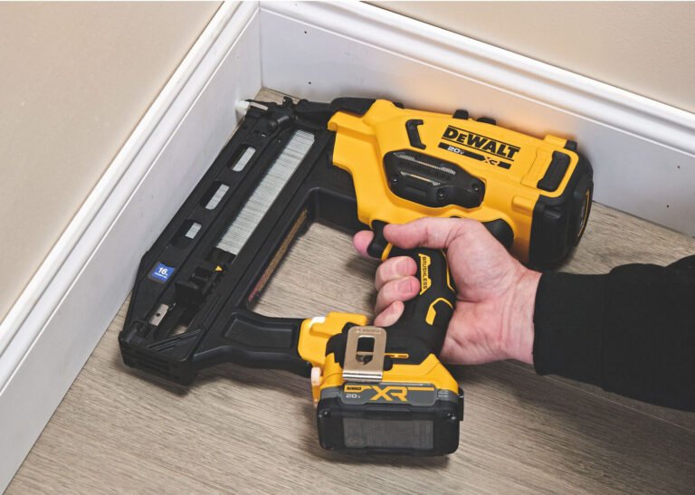 A hand holding a yellow and black DEWALT cordless stapler, being used to secure baseboard trim in a corner on a light-colored floor.